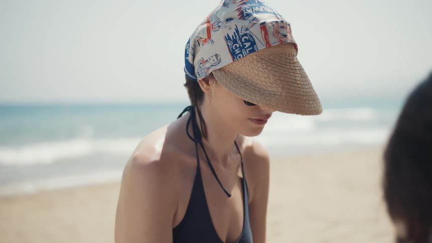 A woman wearing a dark blue swimsuit a straw visor and a bandana on her head stands next to a person in the foreground while on a bright sandy beach