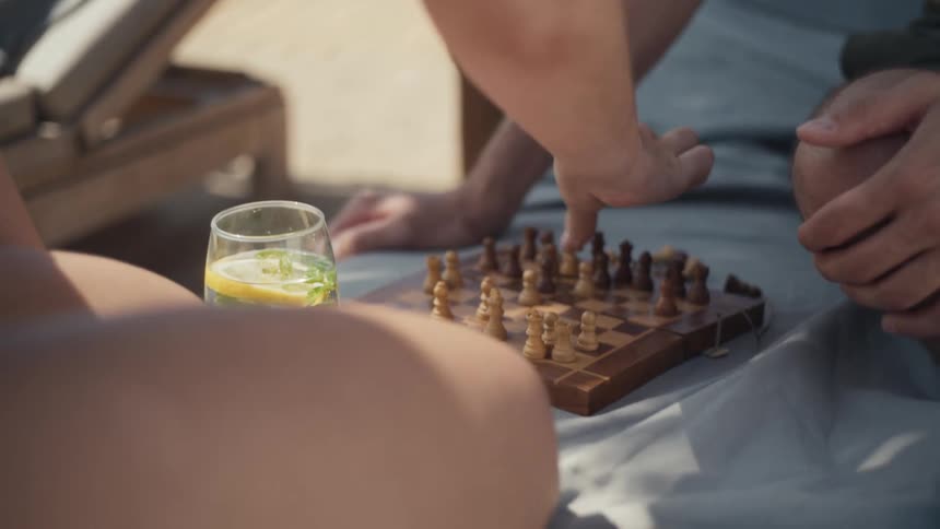 Two people playing chess on a small wooden board near a beach lounge with one person making a move and a glass of a cold drink nearby