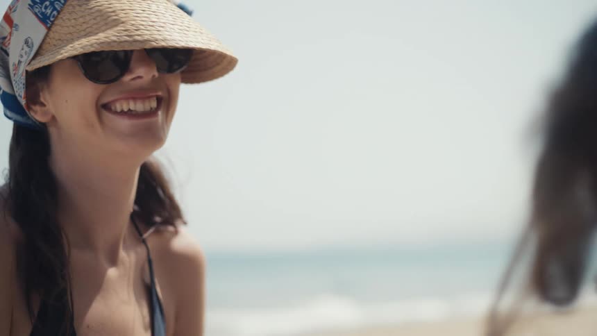 A woman wearing a dark swimsuit and a straw visor looks up and then laughs while talking to a man who is standing in the foreground