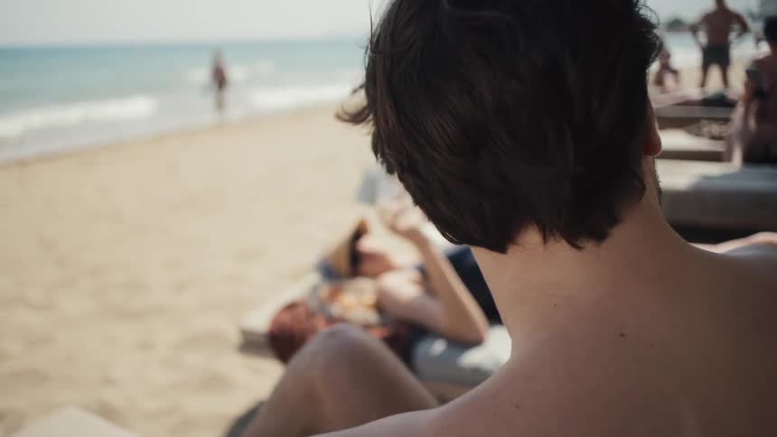 A man with sunglasses and a beard shirtless relaxes on a beach chair taking a sip from a glass while people and waves are visible in the background