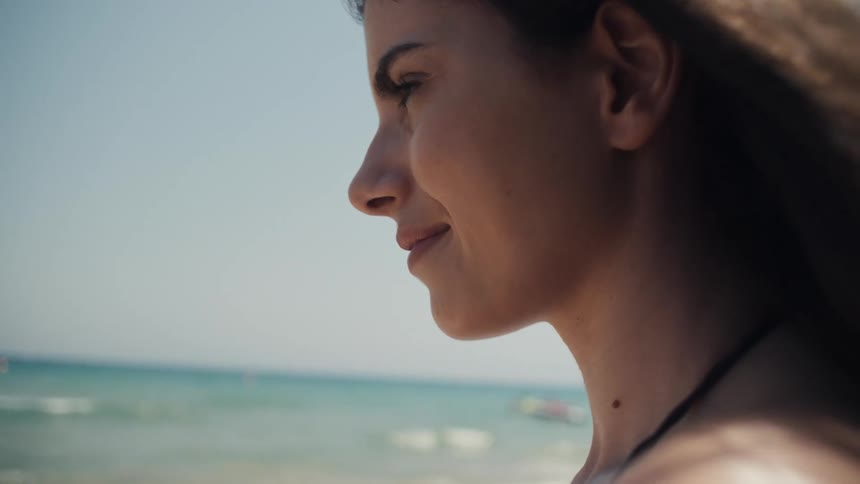 A closeup of a womans face as she stands on a sunny beach she is looking out at the ocean smiling slightly with a relaxed expression