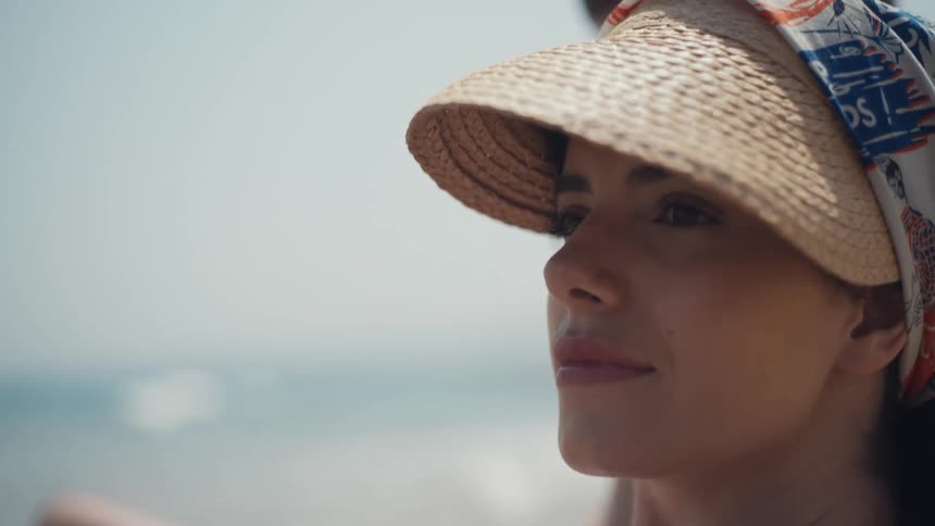 A woman wearing a wide brimmed straw hat with a patterned scarf sits outdoors with her eyes closed as a soft focus ocean background blurs behind her