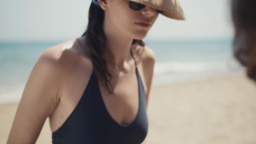 A woman in a swimsuit sunhat and sunglasses concentrates intently on something in front of her while on a sunny beach with a man partially visible in the foreground