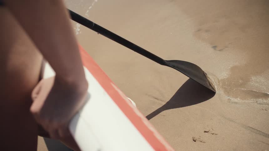 A person holds a black paddle and a white and orange paddleboard at the edge of the ocean where the sand is wet