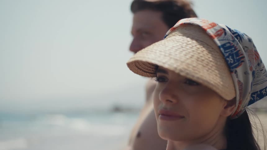 A couple stands on the beach looking out at the ocean the woman wearing a sun visor is in the foreground while the man smiles in the background