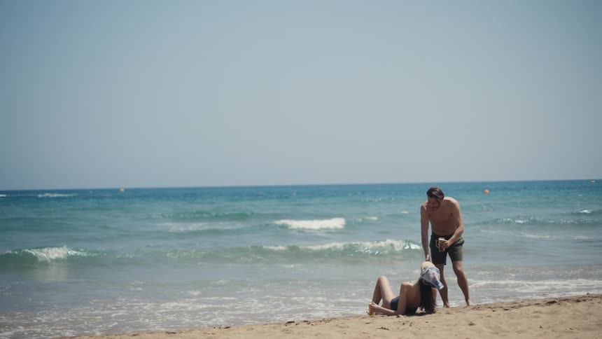 A shirtless man holding drinks walks toward a woman lounging on a chair at the edge of a sunny wavy beach