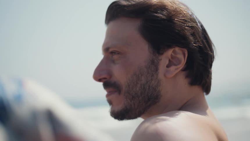 A man with a beard and brown hair smiles as he looks at a woman offscreen while his bare shoulder is visible and the ocean is a blurry background