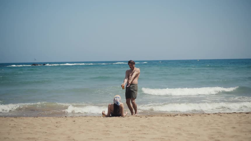 A shirtless man stands and playfully gestures with a drink in his hand while talking to a woman who is sitting in the sand with her back to the camera
