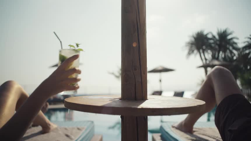 Person holding a cocktail with a straw and mint while relaxing on a poolside lounger