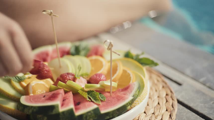 A hand reaches into a white platter of cut fruit including watermelon strawberries and oranges all resting on a wicker placemat beside a swimming pool