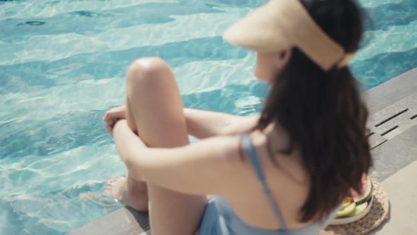 A woman sits on the edge of a swimming pool with her feet in the water next to her is a straw hat and a plate of sliced fruit