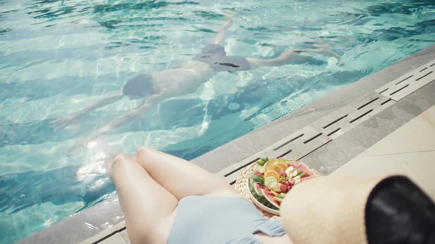 A woman relaxes poolside with a tray of fresh fruit beside her while a man swims underwater in the clear blue pool