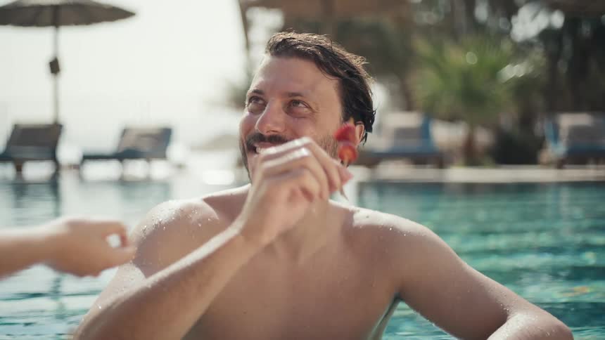 A man is smiling in a swimming pool on a sunny day a womans hand offers him a strawberry skewer which he takes and eats happily