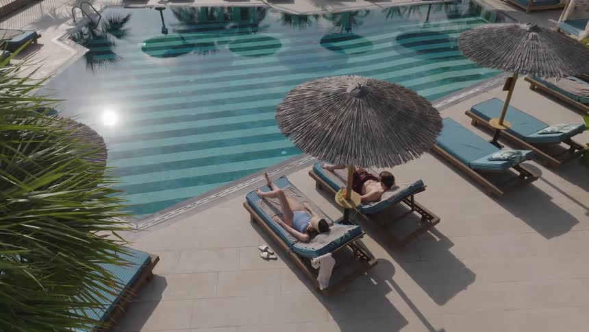 Couple relaxes on poolside loungers under straw umbrellas at a resort