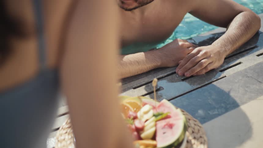 A man is smiling at a woman from inside a swimming pool a plate of fresh sliced fruit is on the poolside and the womans hands reach for a piece
