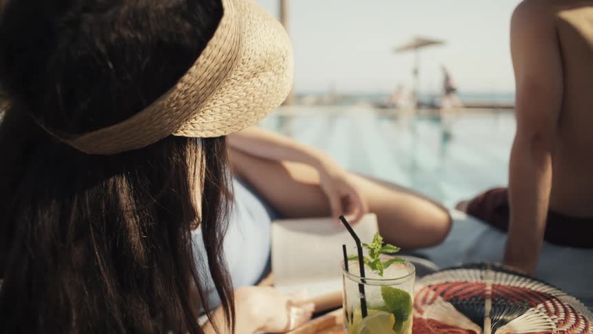 A closeup shows a refreshing cocktail with mint and lime on a wooden tray a person wearing a sun visor and a person with sunglasses are lounging by the pool in the background