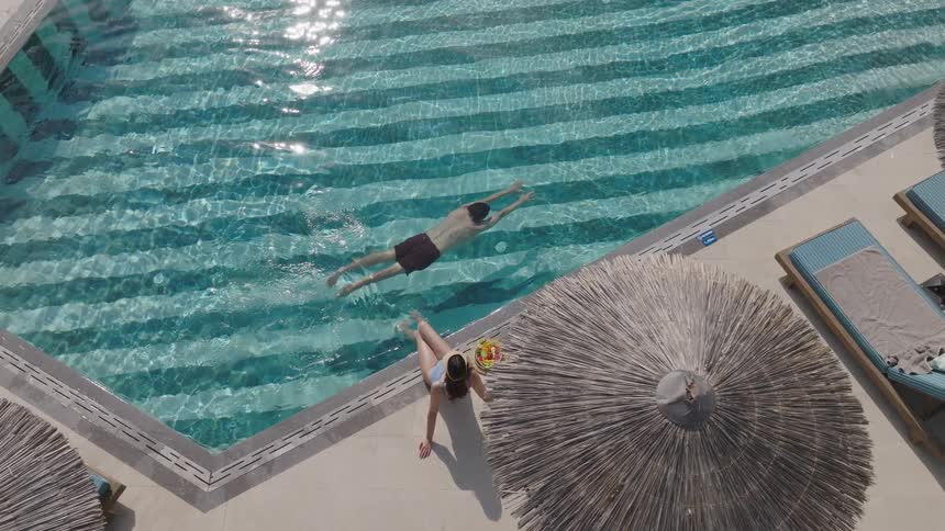 Man swimming in the pool while a woman sits at the edge with a plate of fruit beside her