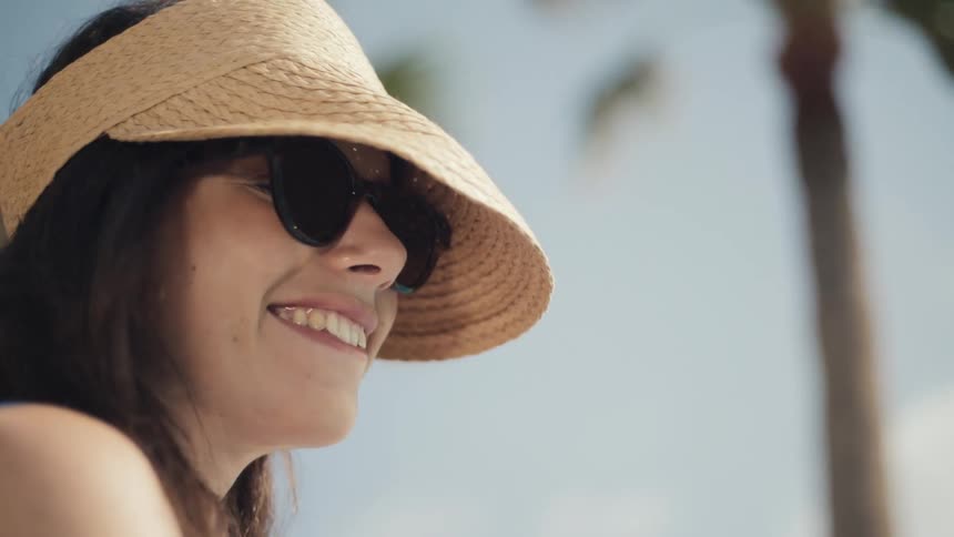 A woman wearing sunglasses and a straw sun visor is smiling and looking offcamera a palm tree is visible in the blurred background suggesting a tropical setting