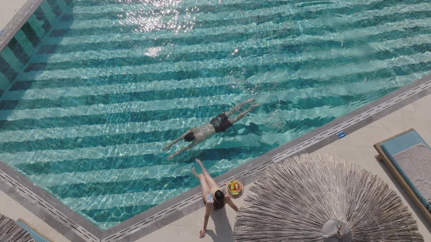 Man swims in the pool while woman in a hat relaxes sitting on the edge with a fruit bowl
