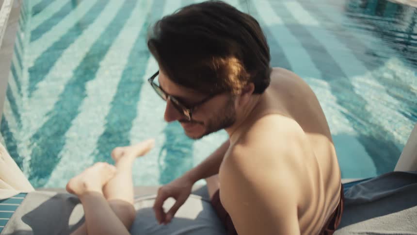 A man and a woman are relaxing by a swimming pool with the man turning to take a drink from a glass while wearing sunglasses