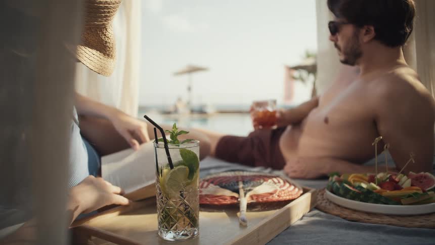 Woman reading a book and man wearing sunglasses relaxing beside drinks and a plate of fruit by the pool