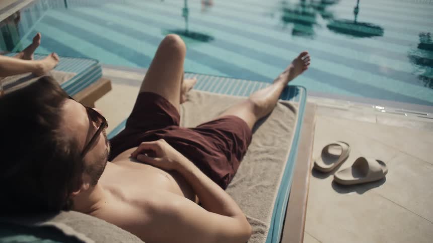 A woman in a blue onepiece swimsuit relaxes on a lounge chair by a pool she is accompanied by a man who is also lounging and wearing sunglasses