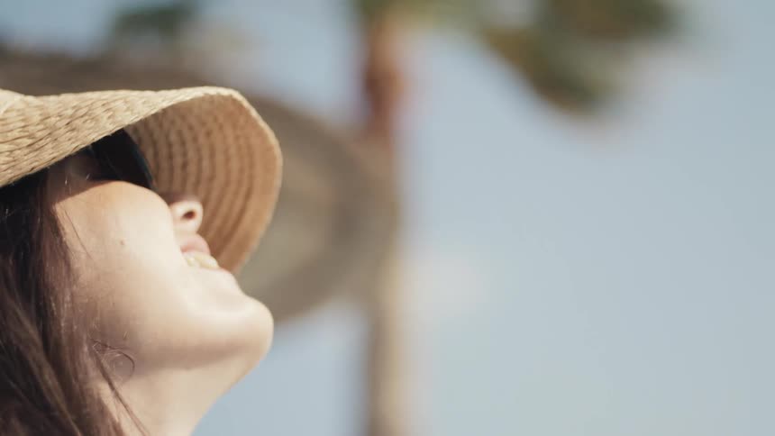 A smiling woman in sunglasses and a straw visor is enjoying the sun she is outdoors with a palm tree in the background