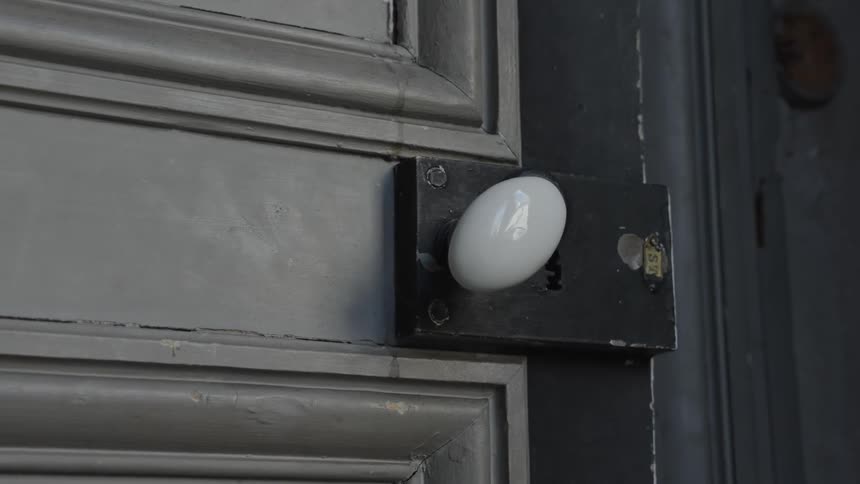 The video shows a closeup of a hand turning a white doorknob on a gray wooden door the camera focuses on the turning of the knob which has a vintage design as the door opens slightly