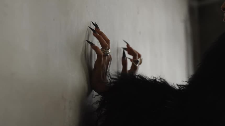 A woman with long nails wearing several rings on her fingers and a black fur coat places her hands on a concrete floor indoors