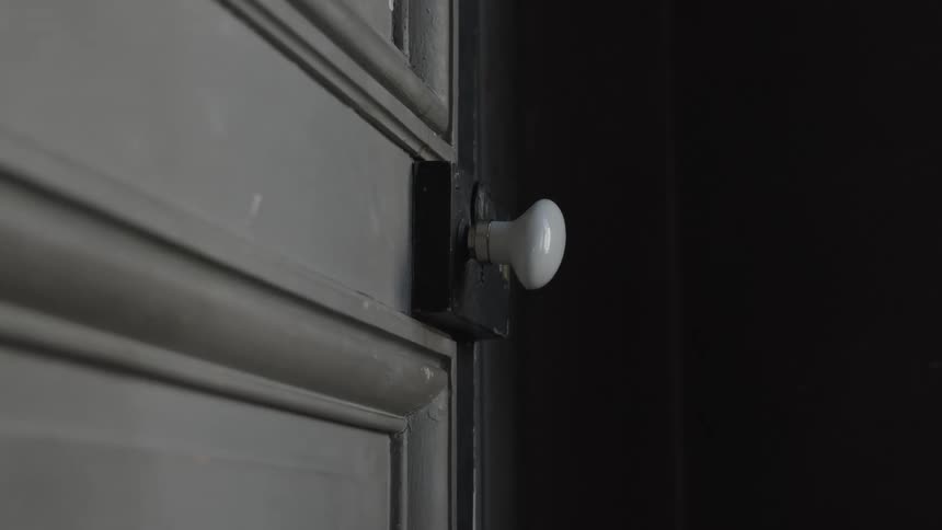A closeup shot shows a persons hand turning a white doorknob the door itself is a darkcolored paneled surface