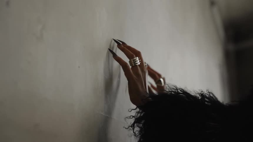 A hand with long black nails and silver rings drags slowly across a wall clad in a black furry sleeve