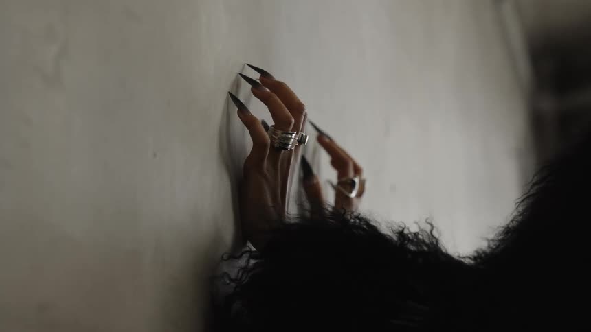 A person with long sharp black fingernails and rings on their fingers is pressing their hands against a wall the person is also wearing a black fur jacket