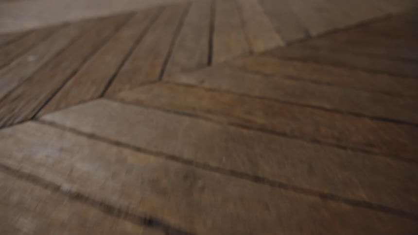 A closeup of a wooden floor shows a worn textured herringbone pattern in soft natural light