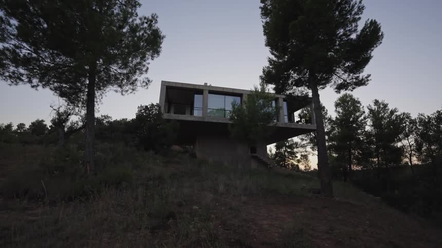 A concrete and glass modernist house with large windows is built on a steep hill surrounded by tall pine trees and a darkening sky overhead