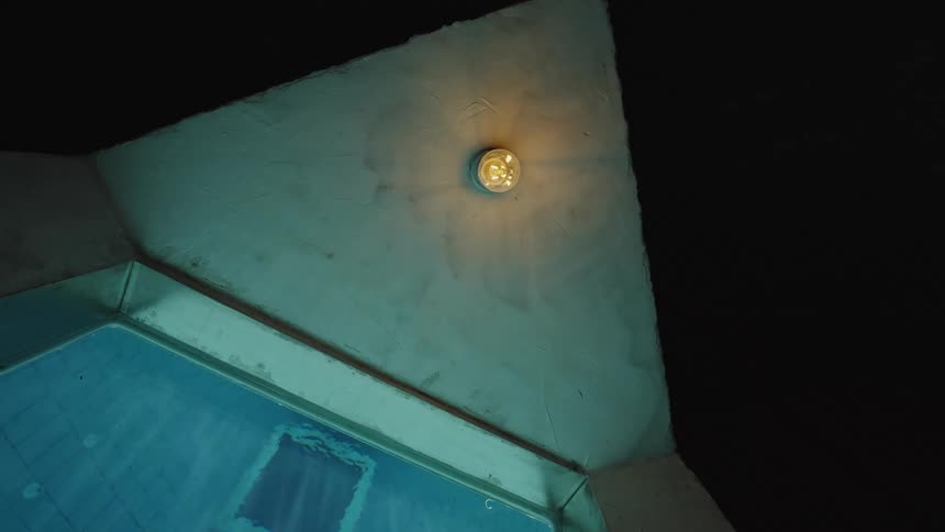 A dim poolside wall glows with a single light fixture above the water at night