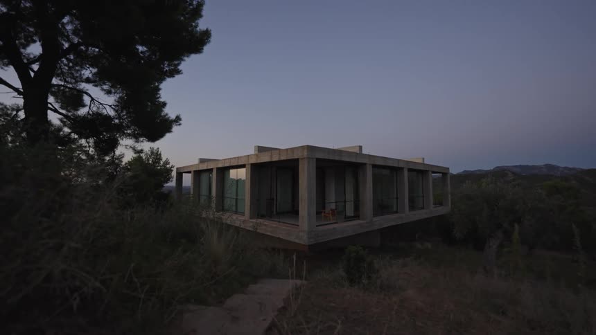 A modern house sits elevated in a natural wooded landscape at dusk