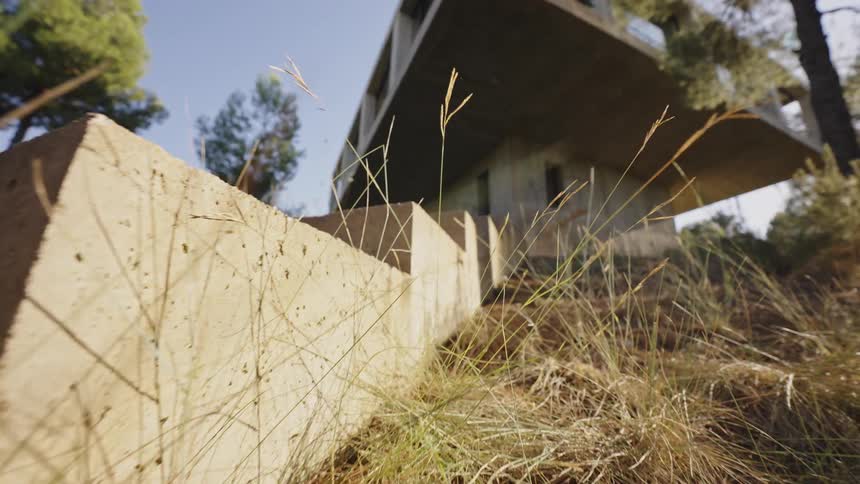 The video shows an exterior shot of a modern house built with concrete the camera pans upwards from a dry grassy area revealing the brutalist architecture of the building