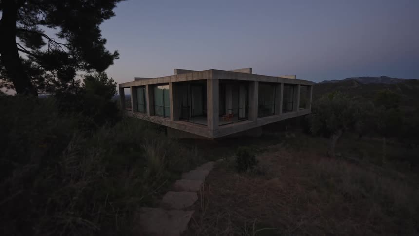A concrete and glass modernist house is nestled on a hillside covered with tall grass with a stone path leading up to it as the sky darkens in the background