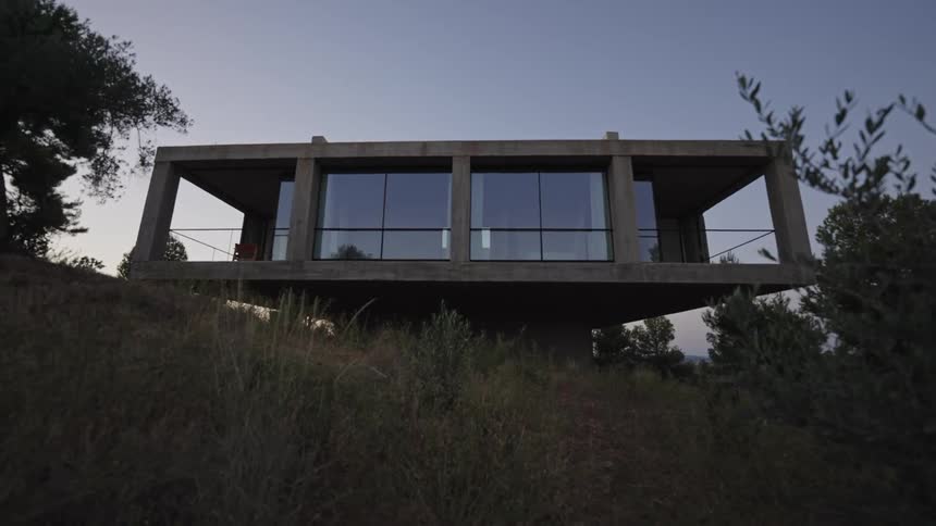 A lowangle shot shows a modernstyle house with large windows the house is situated on a grassy hill surrounded by trees