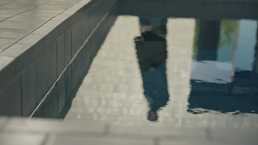 Reflection of a standing person appears in the water of a tiled pool