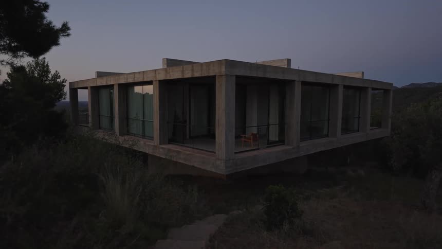 A modern concrete house with glass walls stands elevated on a hillside surrounded by brush at dusk