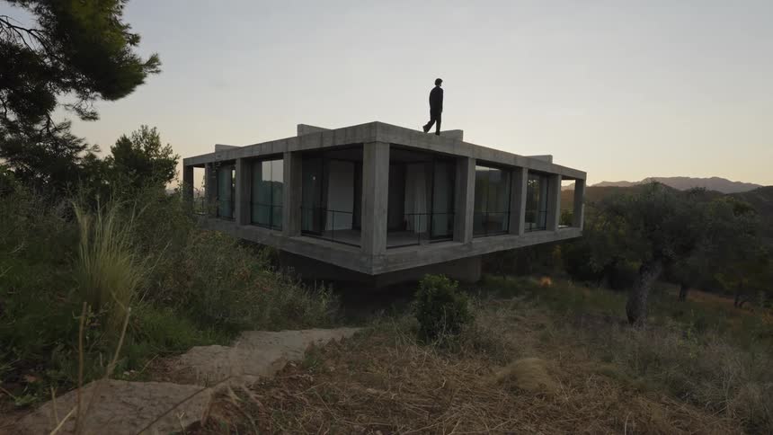 A man walks in silhouette on the flat roof of a modern concrete building with large windows looking out at a hilly landscape under a dusky sky