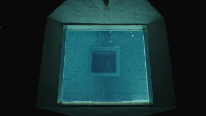A single lightbulb hangs over a large trapezoidal window which looks into a dark swimming pool with blue tiles on the walls and floor