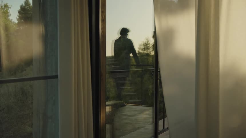 A shot from inside a room shows a persons silhouette standing on a balcony with glass doors and curtains framing the view of trees in the background