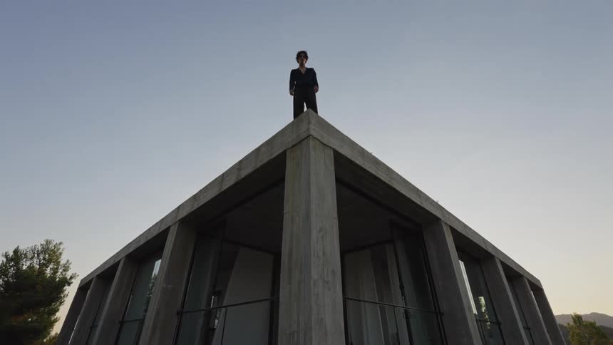 A modern triangular brutaliststyle house with a concrete exterior and large windows is shown with a person standing majestically on its highest point against a clear sky