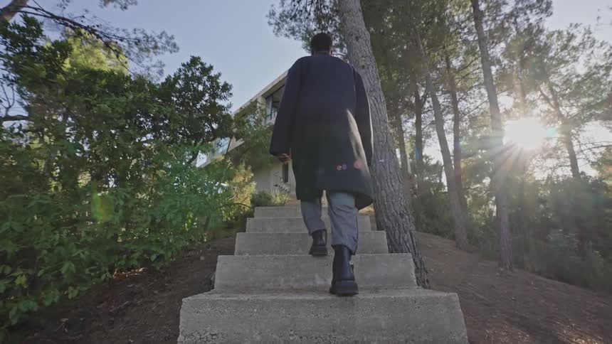 Person in a black overcoat walking up concrete stairs towards a modern house surrounded by trees