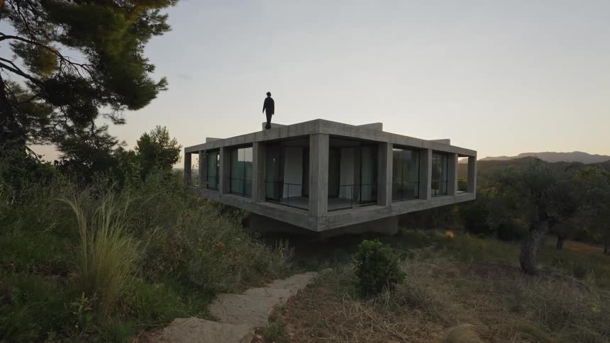 A man walks in silhouette on the flat roof of a modern concrete building with large windows looking out at a hilly landscape under a dusky sky007