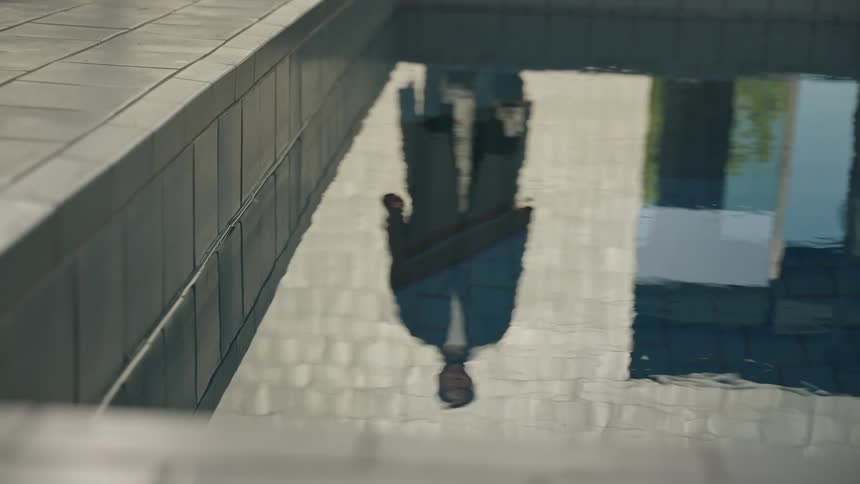 A distorted reflection of a person appears in the calm tiled water beside a modern pool