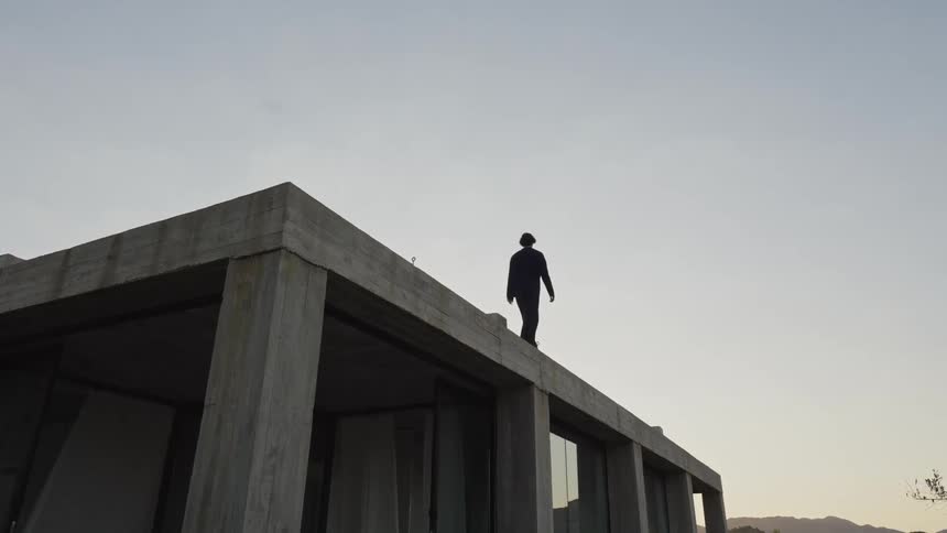 A lowangle shot shows a man standing on the roof of a concrete building the man is silhouetted against a pale sky as the camera slowly zooms out