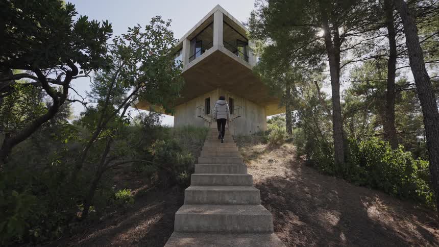 A person in a puffer jacket climbs stone steps toward a modern elevated house nestled in the forest